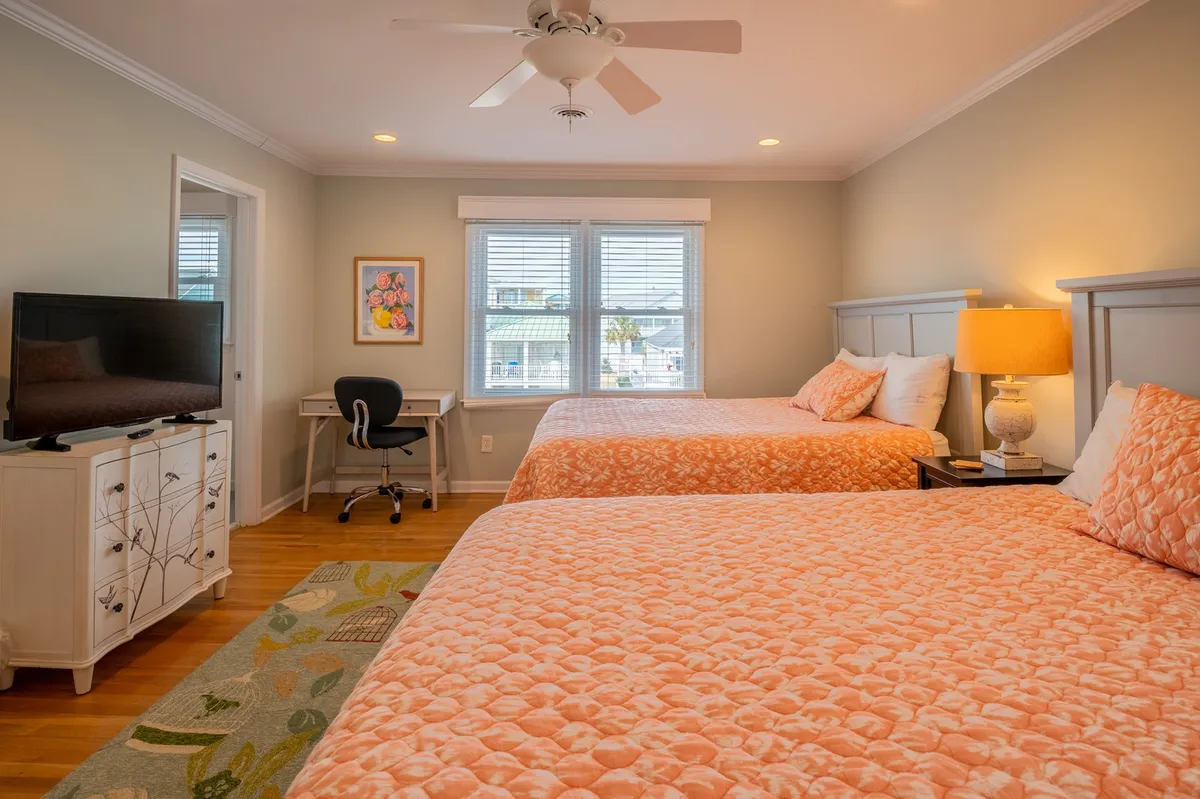 A comfortable bedroom with two beds adorned in coral-colored bedding, a small desk and chair for work or study, a TV on a white dresser, and cheerful wall art. Sunlight filters through the windows, creating a cozy space ideal for relaxing, watching TV, or making memories with family.