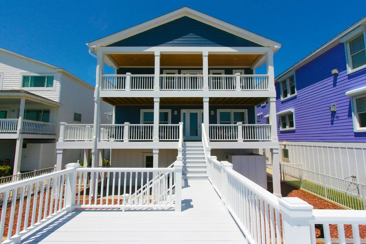 The exterior hero image showcases a striking blue beach house with a wide, inviting white staircase leading to expansive balconies lined with classic railings. Sunlight bathes the facade, while the spacious decks are ready for gatherings, morning coffee, and breathtaking ocean views. The scene radiates warmth and the promise of joyful seaside living, with guests pictured relaxing and sharing moments on the porches.