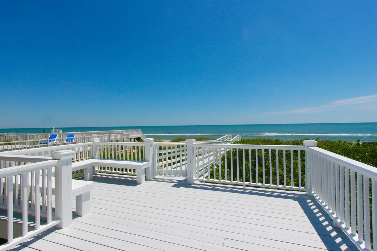 A wide deck with direct beach access, offering clear views of the ocean and sandy dunes. The deck space is open, inviting sunbathing, reading, or group gatherings, and it emphasizes an easy transition from the home to the shore.