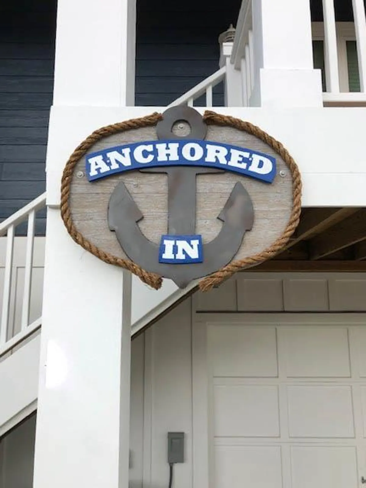 An exterior scene featuring a sign with 'Anchored In,' accented by nautical rope and an anchor design. The setting suggests a friendly, welcoming arrival experience, with hints of sunset light and the social energy of friends and family gathering nearby. It hints at the unique identity and sense of place the property offers.