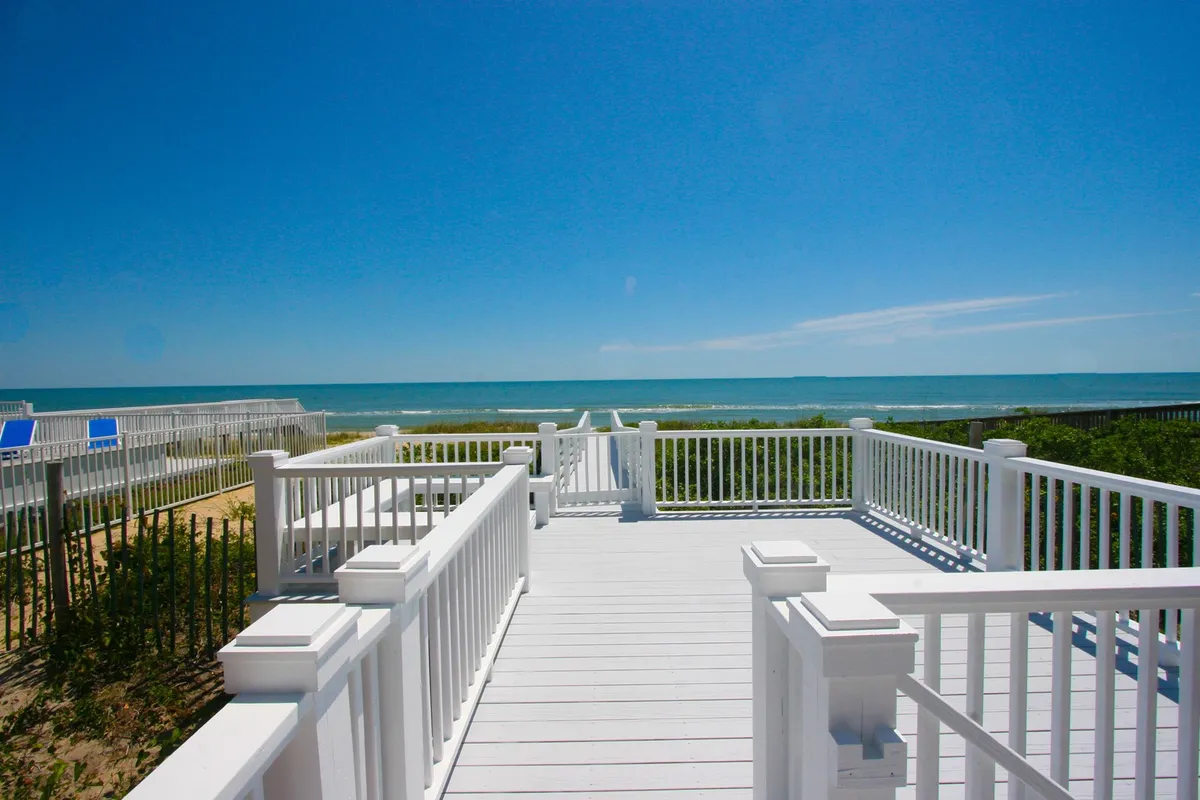 This image features an exterior view from the home's deck, overlooking the ocean with visible sand dunes and a sturdy deck railing. The angle captures the direct connection from the house to the sweeping beach vistas, while the deck itself invites thoughts of sunbathing, outdoor dining, or simply relaxing while enjoying the coastal breeze.