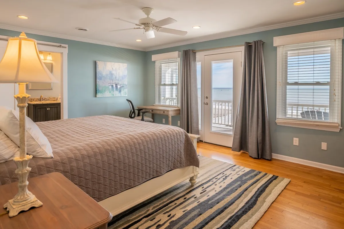 A serene bedroom with a cozy bed positioned next to a large window offering a glimpse of the ocean. The room includes a desk, a lamp, and a painting, creating a peaceful retreat for relaxation, reading, or quiet reflection. The window showcases natural light streaming in, hinting at the beautiful seascape views available from the property.