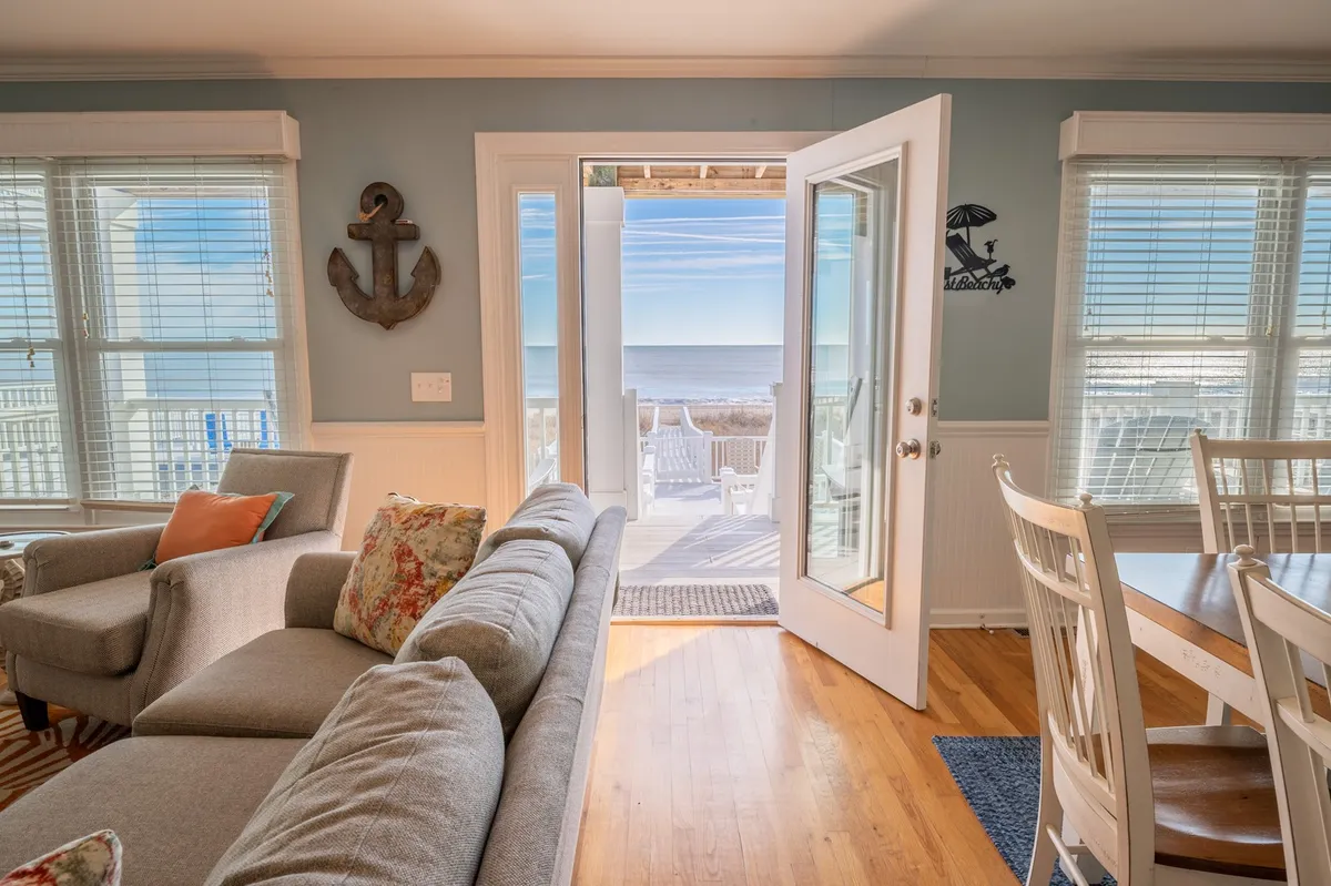 This image features an open door leading directly out to the beach, with a comfortable sofa and a dining table visible in the foreground. The scene is bright and airy, suggesting easy access to outdoor adventures and gathering spots for families and friends.