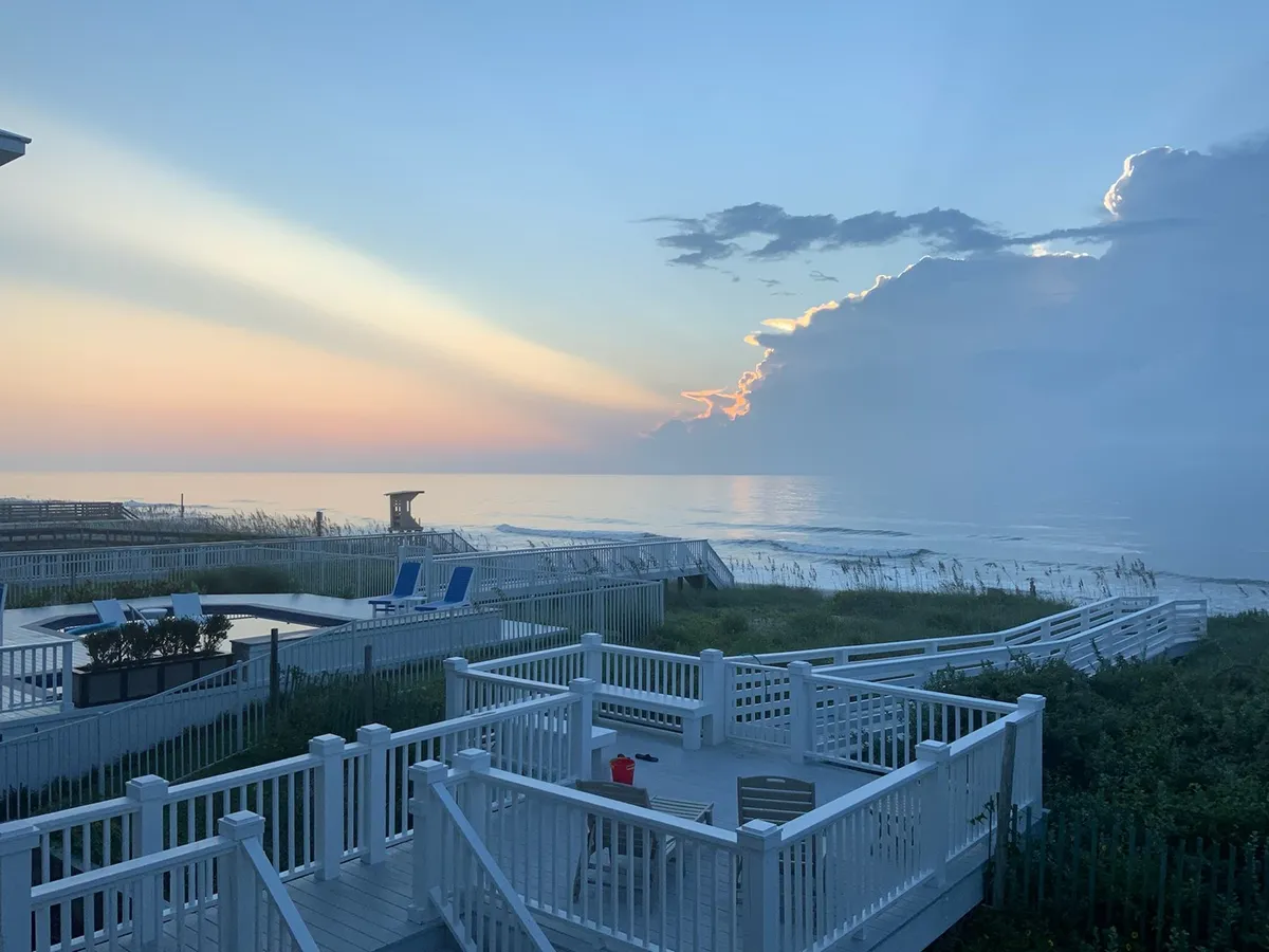 A serene exterior shot highlighting the home's expansive deck area, complete with a pool in the foreground, ocean waves in the background, and the sun setting on the horizon. This inviting perspective suggests the joys of relaxing by the pool, enjoying direct ocean access, and gathering outdoors as day turns to night.