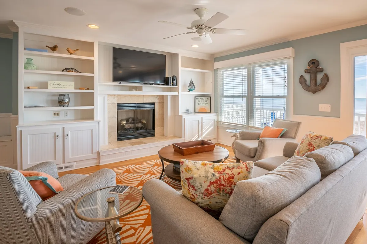 This image features a cozy great room with a fireplace, large windows that let in abundant natural light, a television, a colorful rug, and plush, comfortable seating. The space feels open and inviting—ideal for relaxing or gathering for movie nights.