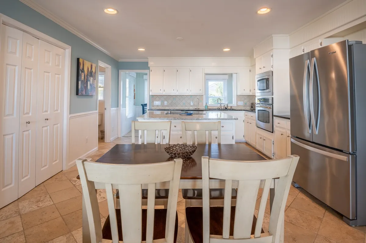A dining area adjacent to the kitchen, with a large table set for family gatherings and artwork on the wall. Kitchen appliances are visible in the background, underscoring the ease of moving from meal prep to shared dining, and the space is styled for entertaining guests or enjoying game nights.
