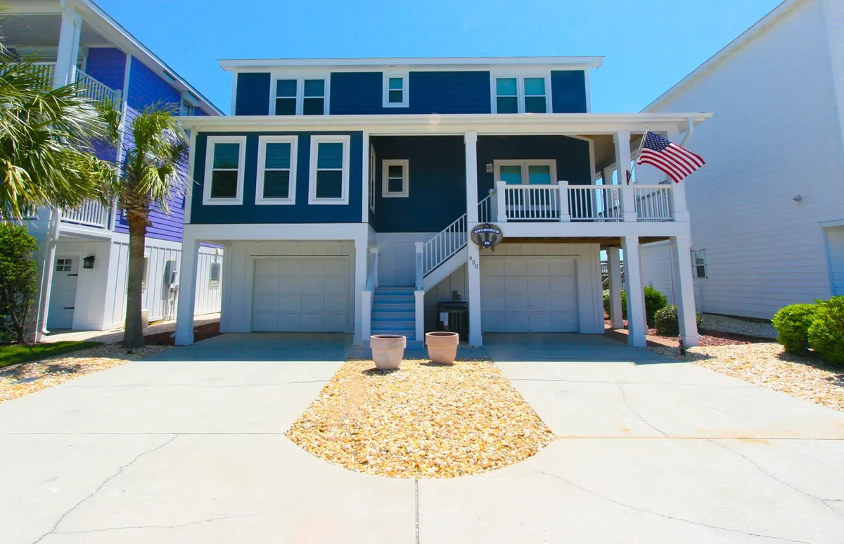 This image shows the cheerful blue facade of the house, white railings, an American flag, and a spacious pebbled driveway. The driveway is clearly visible and able to accommodate multiple vehicles, directly highlighting the 'ample parking' amenity mentioned in the section. The welcoming exterior suggests ease of arrival and electronic entry.